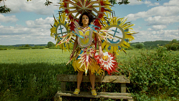 Film still of Rhea Storr's 'A Protest, A Celebration, A Mixed Message' depicting a woman wearing a large, colorful Carnival costume while standing on a wooden bench in front of a green field.