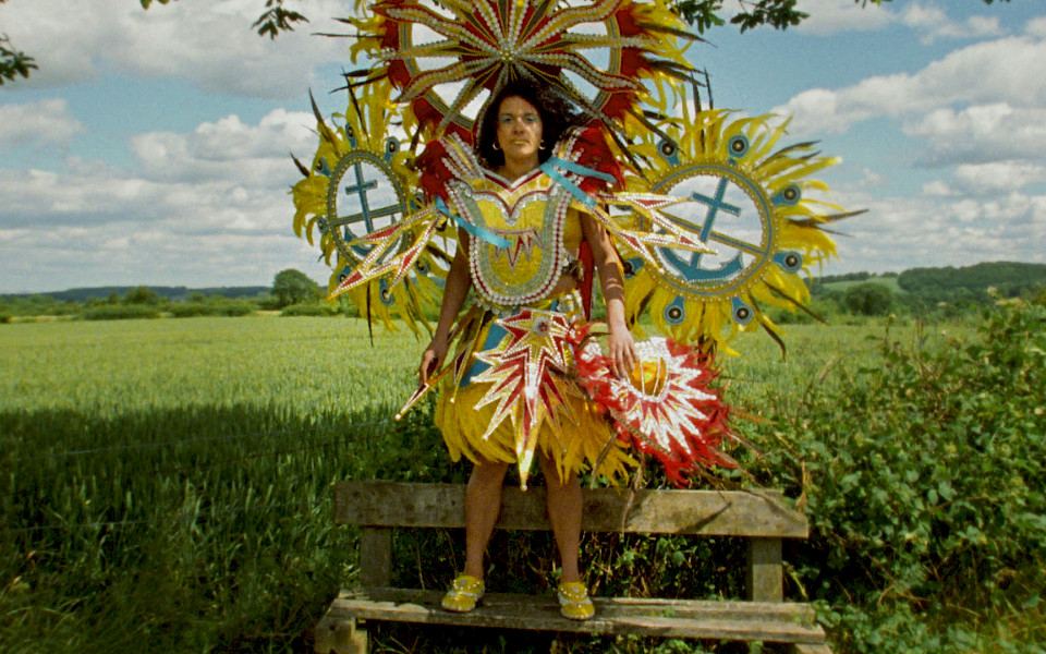 Film still of Rhea Storr's 'A Protest, A Celebration, A Mixed Message' depicting a woman wearing a large, colorful Carnival costume while standing on a wooden bench in front of a green field.