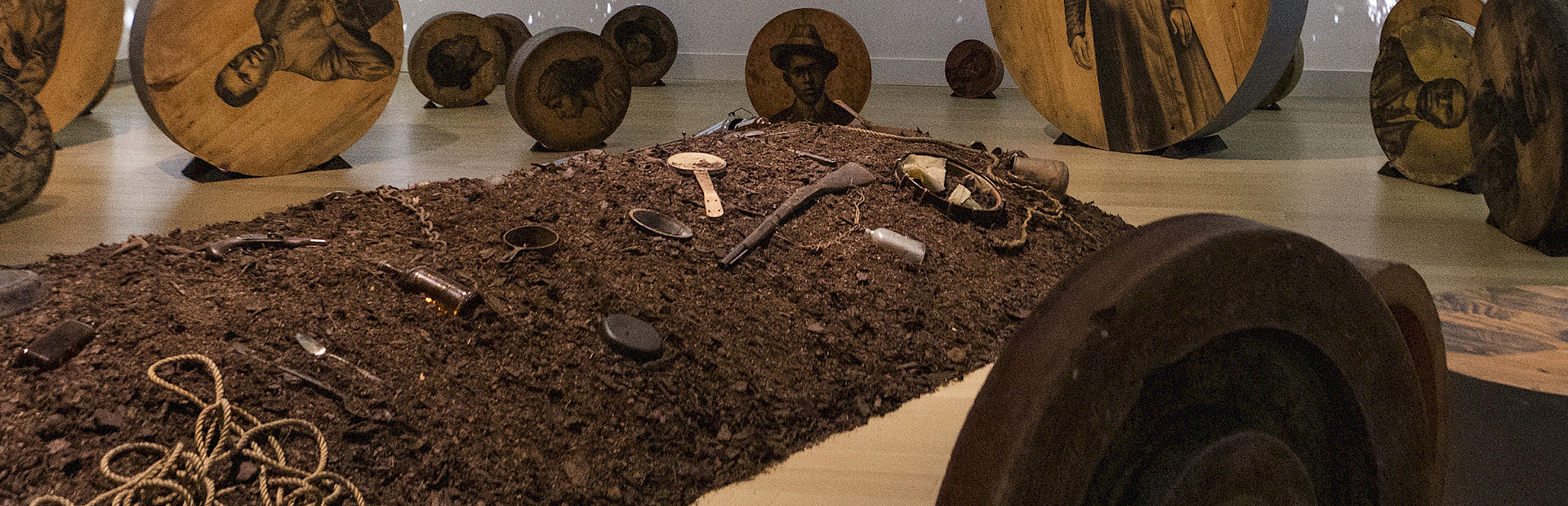 Photo of an artwork installation featuring a mound of dirt on the floor covered in objects surrounded by charcoal drawings of people on large round pieces of wood with a video of water projected on the walls behind them.