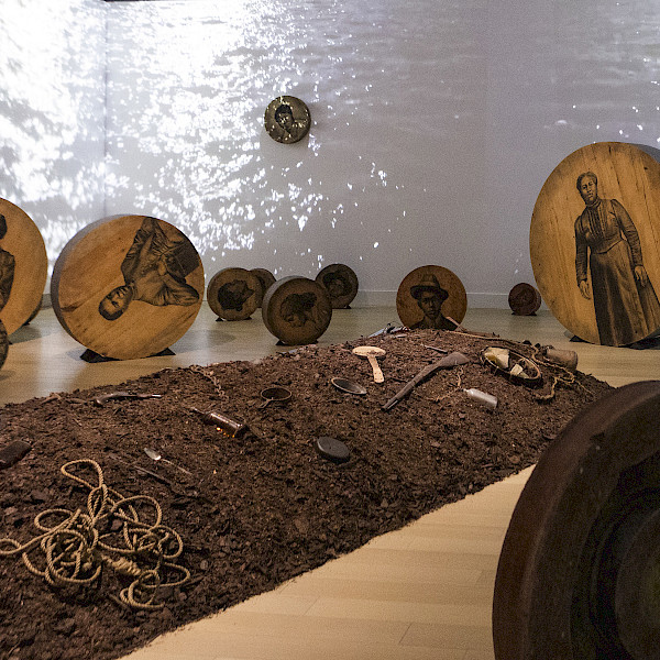 Photo of an artwork installation featuring a mound of dirt on the floor covered in objects surrounded by charcoal drawings of people on large round pieces of wood with a video of water projected on the walls behind them.