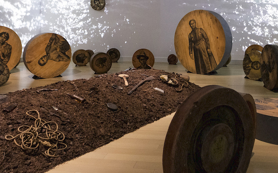 Photo of an artwork installation featuring a mound of dirt on the floor covered in objects surrounded by charcoal drawings of people on large round pieces of wood with a video of water projected on the walls behind them.