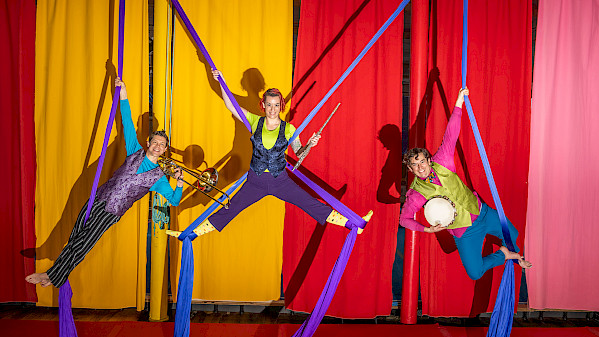 Photo of a trumpet player, flute player, and tambourine player hanging from purple silks above a stage with red and yellow curtains behind them.