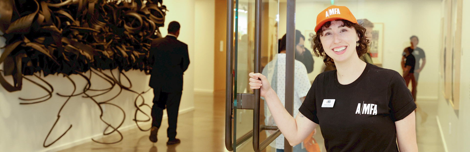 Photo of a woman wearing an AMFA T-shirt and hat holding open the door to an art gallery.