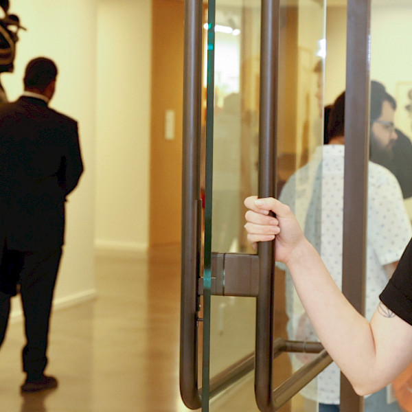 Photo of a woman wearing an AMFA T-shirt and hat holding open the door to an art gallery.