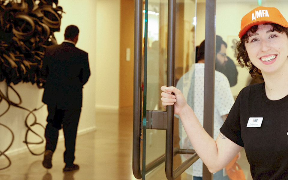 Photo of a woman wearing an AMFA T-shirt and hat holding open the door to an art gallery.