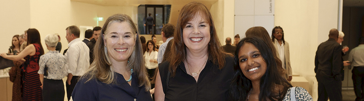 Photo of three women smiling in the Atrium of AMFA surrounded by other people during an event.