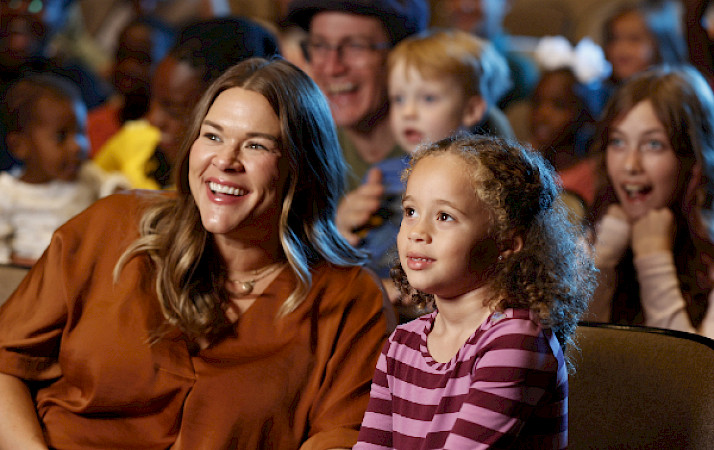 Photo of a woman and a young girl seated and smiling in a theater with other people seated around them.
