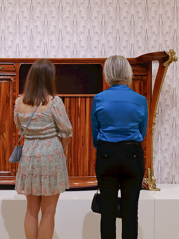 Photo of two women looking at Art Nouveau furniture in a gallery at the Arkansas Museum of Fine Arts.