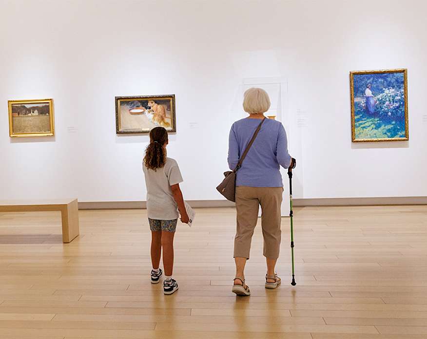 Photo of a young girl and elderly woman walking with a cane looking at art in an art gallery.