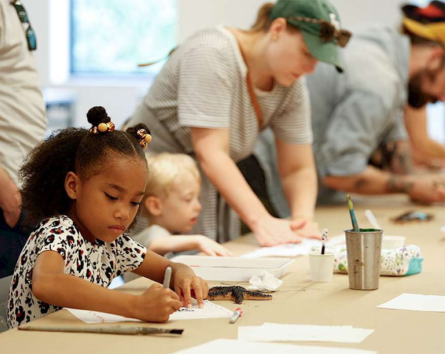Photo of a young girl drawing on paper on a table in an art studio. Other children and adults are doing the same in the background.