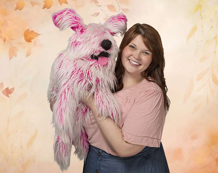 Photo of a woman wearing a pink shirt holding a pink dog puppet in front of a leafy background.