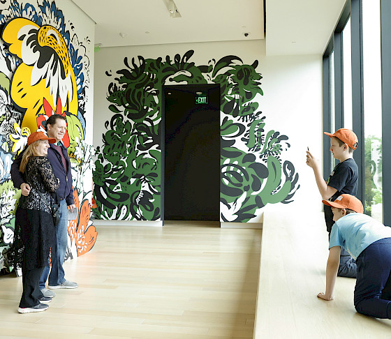 Photo of two children taking a photo on a cellphone of their parents posed in front of an art installation.