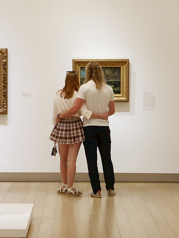 Photo of a couple with their armas around each other looking at a painting in an art gallery.