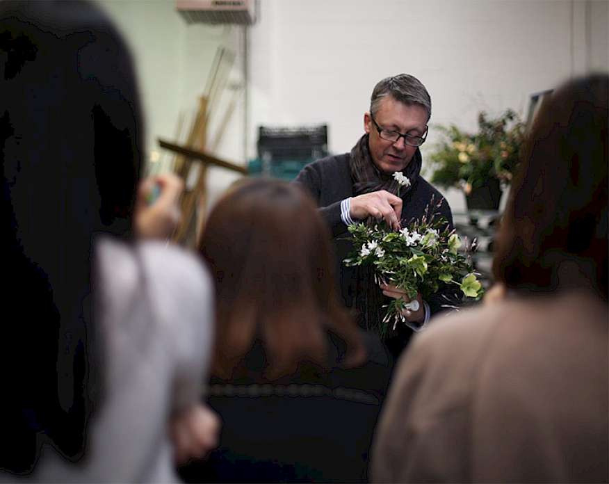 Photo of a man with short gray hair wearing glasses and a dark sweater and scarf arranging flowers in front of a group of people.