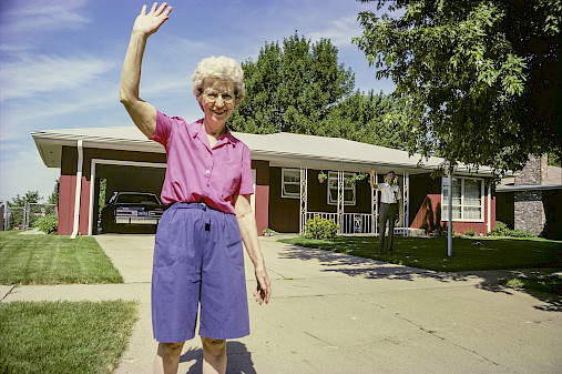 Photo of a woman standing in the driveway of a red house waving.