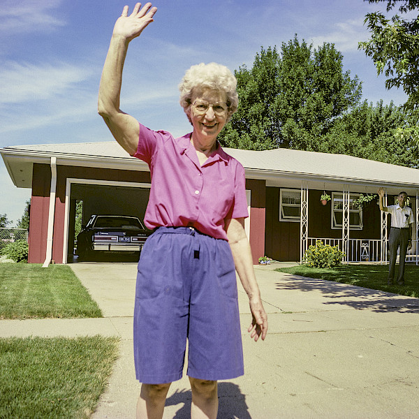 Photo of a woman standing in the driveway of a red house waving.