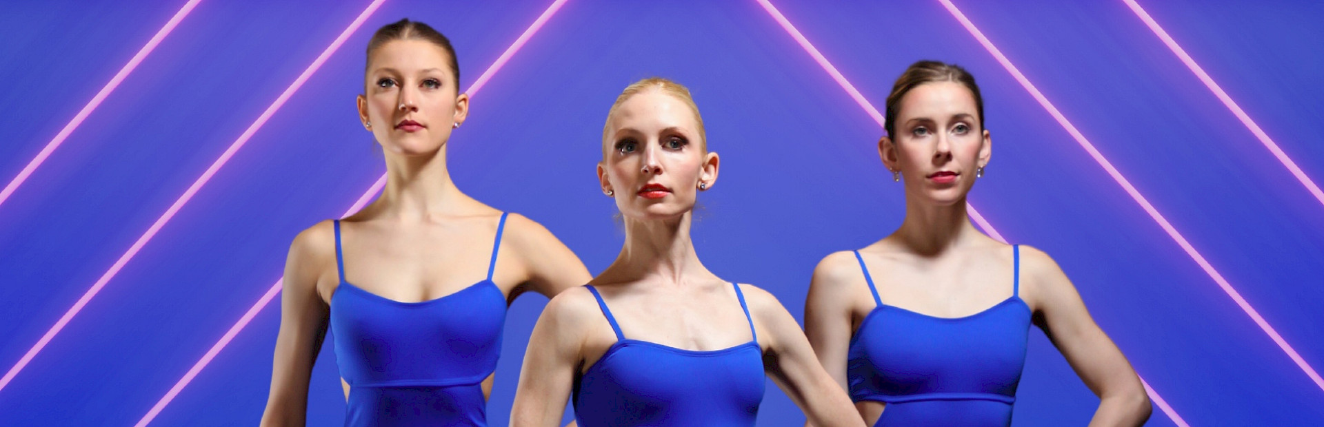 alt="Photo of three ballerinas wearing blue leotards posed in front of a blue background with neon lights."