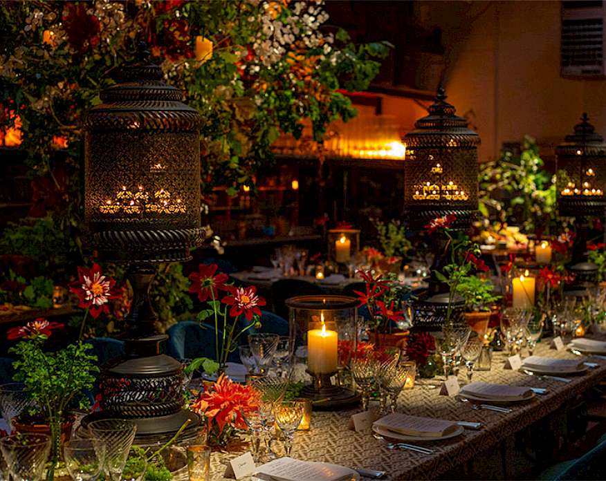 Photo of a long table set with plates, silverware, and glasses next to large floral arrangements and lanterns holding candles.
