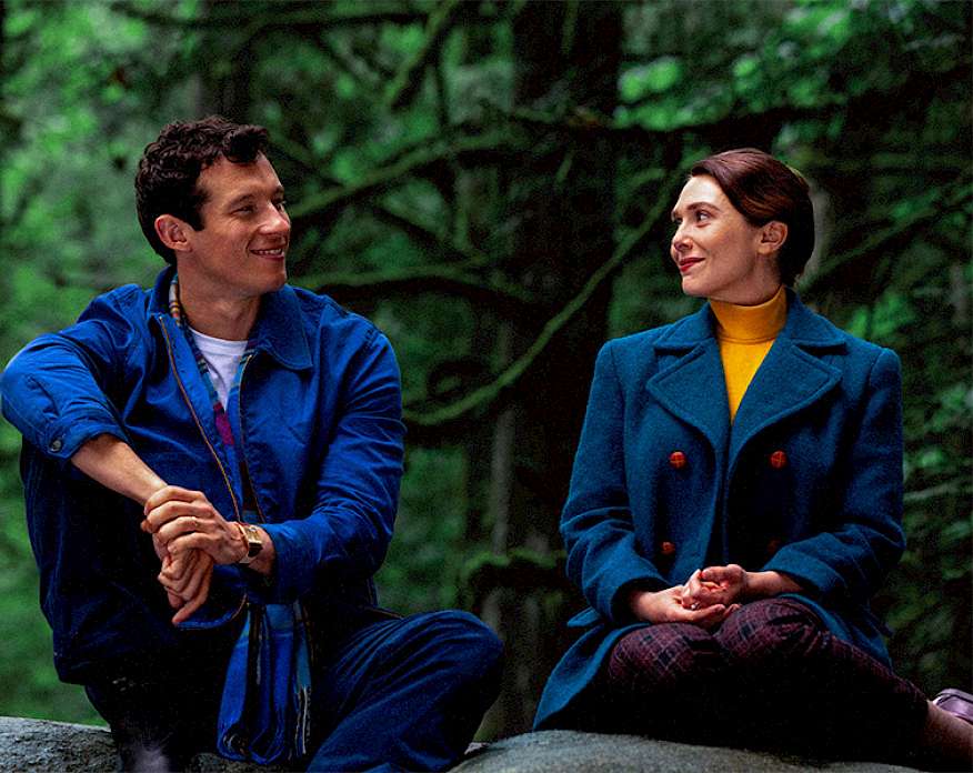 Film still depicting a man and a woman wearing jackets and sitting on a large rock in a forest.