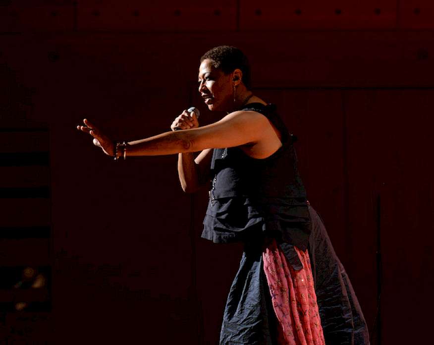 Photo of Lisa Fischer on stage under a spotlight and holding a microphone with her left arm outstretched toward the audience.