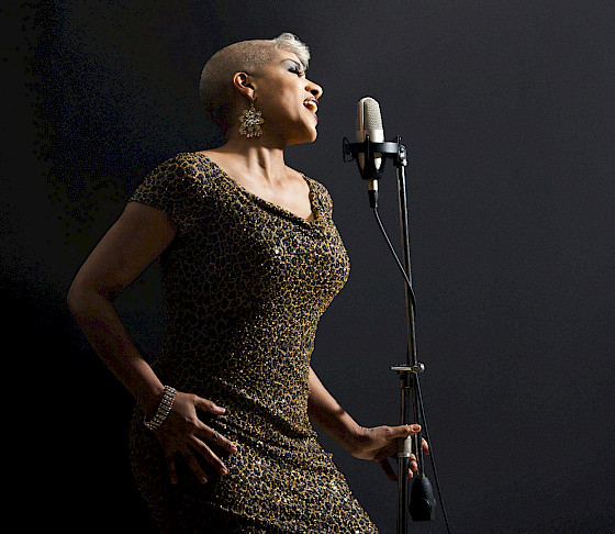 Photo of a woman with short light hair wearing a leopard print dress and dangly earrings singing into a microphone.